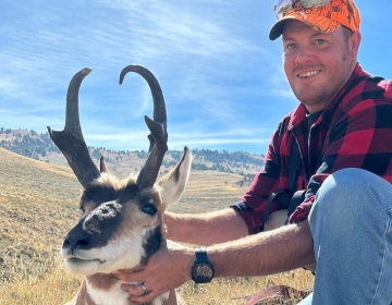 A smiling hunter in a red flannel shirt poses with a pronghorn antelope in the rolling hills of Wyoming. The photo captures the beauty of the terrain and a clear autumn day.